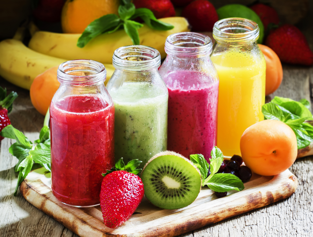 Four glasses of fresh juice on a wooden table with fruits and vegetables in the background 